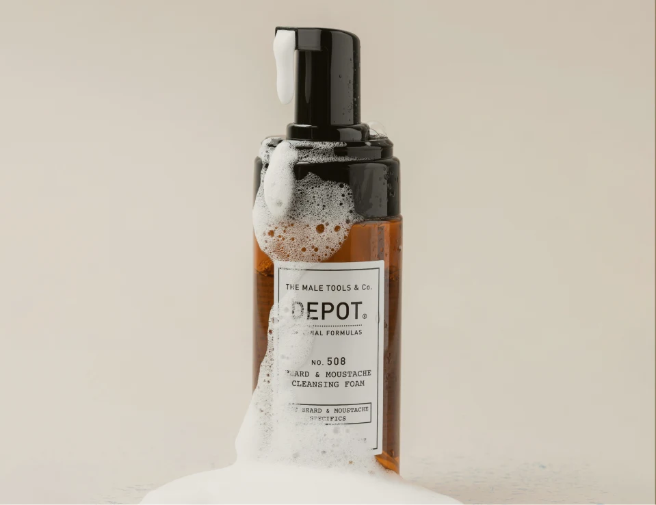 Her ses DEPOT No. 508 Beard & Moustache Cleansing Foam i en transparent brun flaske med sort pumpe og hvidt skum omkring flasken.