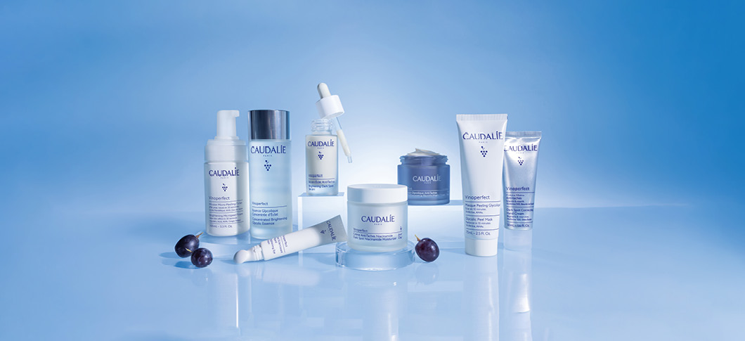 Her ses CAUDALIE hudplejeprodukter i lyse flasker og krukker på en blå baggrund med druer.