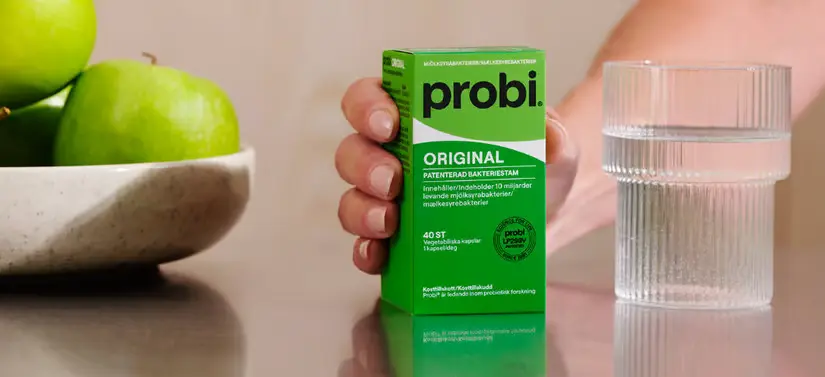 Her ses et bord med en skål æbler i venstre side og et glas vand i højre. I midten er der en hånd, som holder et Probi produkt.