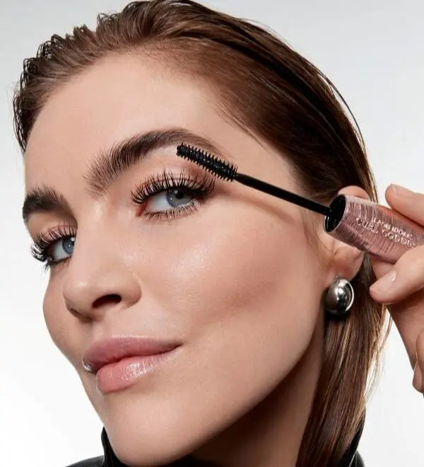 Nærbillede af en kvinde, der påfører mascara på sine øverste øjenvipper med en sort mascarabørste. Hun har markerede bryn, lange vipper og naturlig makeup med glødende hud og lyserøde læber. Kvinden bærer sølvøreringe og ser let opad, mens hun holder mascaraen tæt ved øjet. Billedet har et elegant og moderne beautyudtryk med fokus på øjenvipper og makeup.