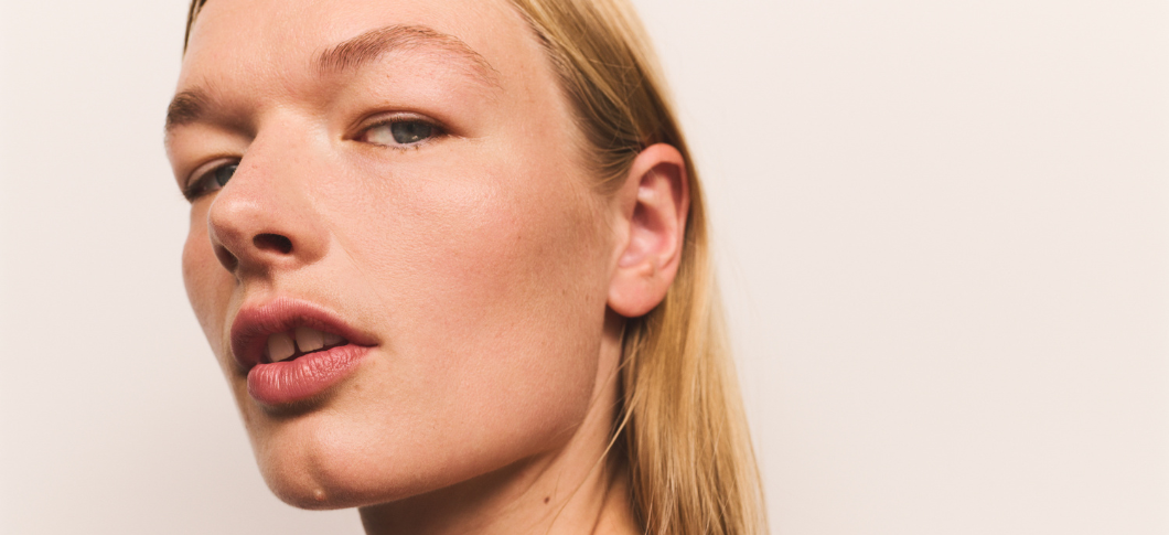 Her ses et nærbillede af en person mod en lys, neutral baggrund. Personen er fotograferet fra siden med fokus på ansigtet, herunder kind, læber og øje. Personen har let makeup.