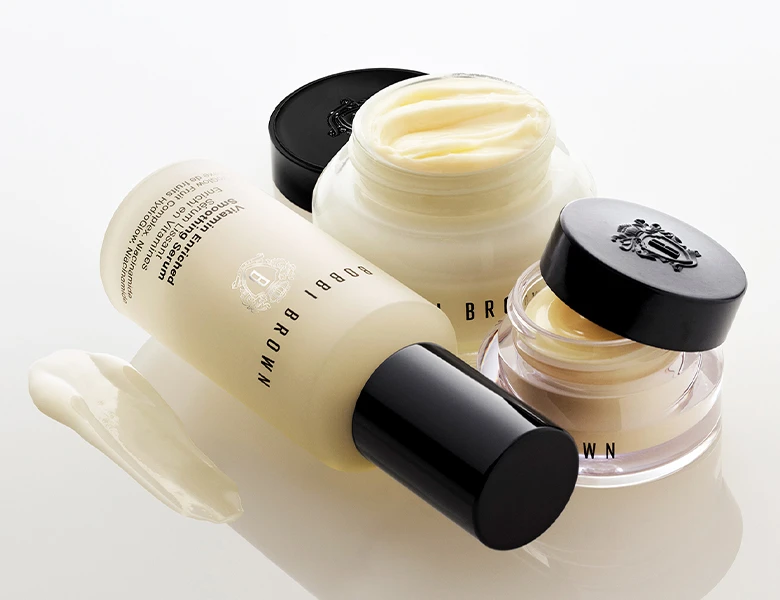 Her ses et billede med hudplejeprodukter fra Bobbi Brown mod en lys baggrund. Produkterne omfatter blandt andet en Vitamin Enriched Face Base, en Vitamin Enriched Face Base Eye Cream og en lotion. Krukkerne er åbne, så den cremede tekstur er synlig, og en lille mængde creme er påført ved siden af produkterne for at fremhæve konsistensen.