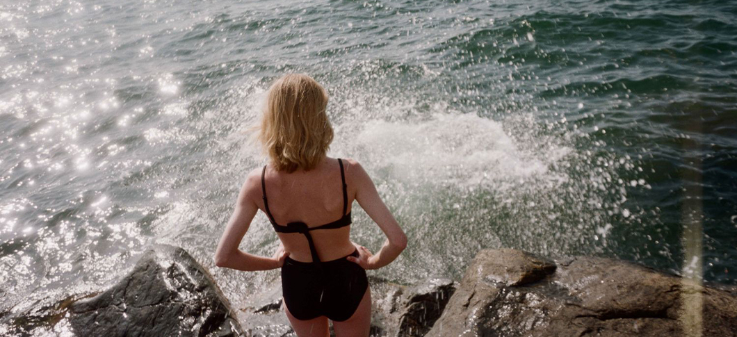 Her ses en person i badetøj stående ved havet med bølger og klipper i baggrunden.