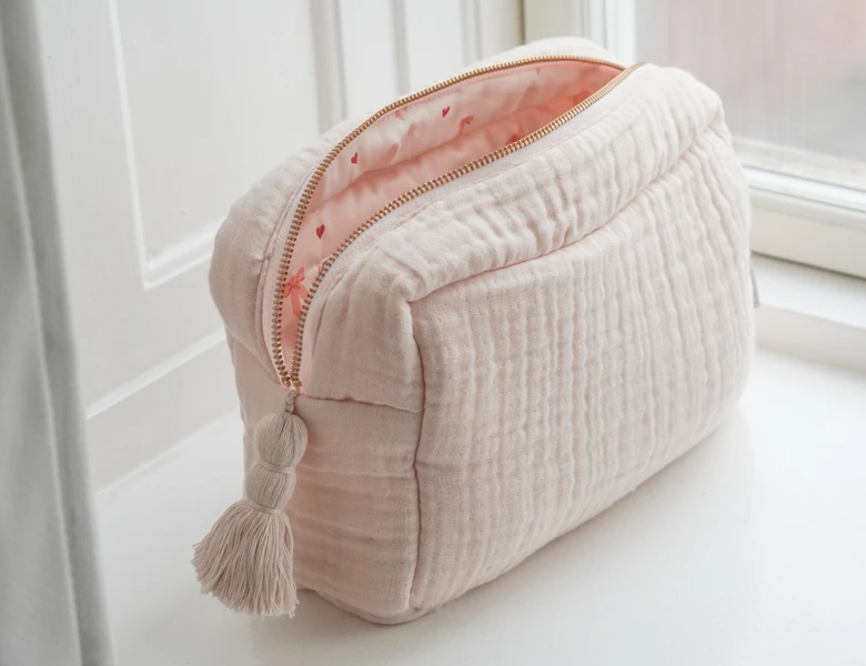 Lyserød quiltet toilettaske med kvast står på en vindueskarm med lynlåsen åben.