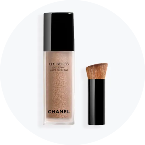Her ses Chanel Les Beiges Eau de Teint Water-Fresh Tint i en transparent flaske med sort låg sammen med en tilhørende makeupbørste.