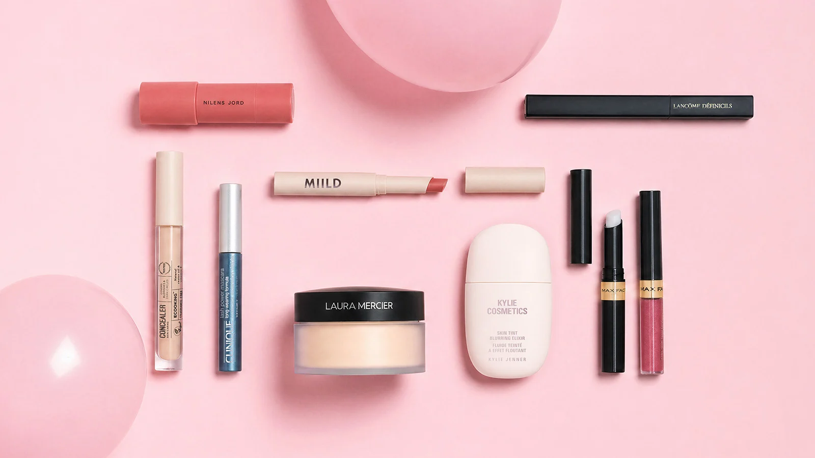 Her ses et udvalg af makeup fra Nilens Jord, Lancôme, Milk Makeup, Ecooking, Clinique, Laura Mercier, Kylie Cosmetics og Max Factor. Produkterne omfatter blandt andet concealer, mascara, læbeprodukter, pudder og skin tint, arrangeret mod en rosa baggrund.