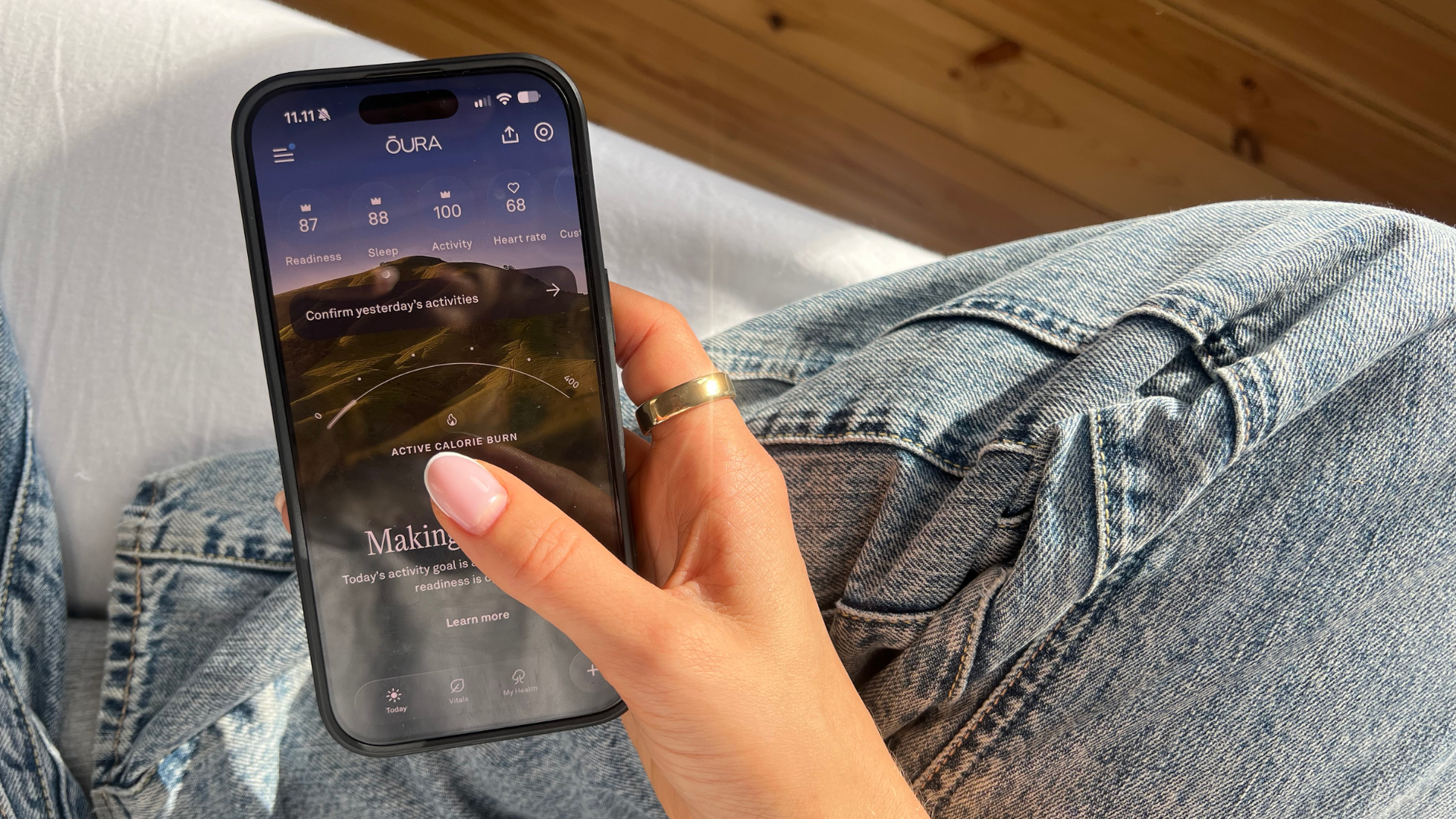 Hånd holder smartphone med Oura-app, der viser sundhedsdata som parathed, søvn, aktivitet, puls og kalorieforbrænding.