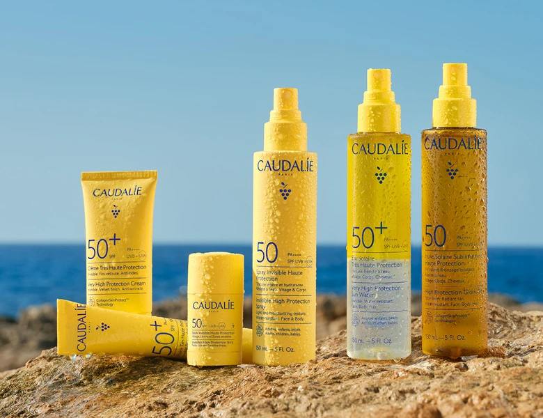 Her ses CAUDALIE solprodukter i gule flasker placeret på klipper ved havet.