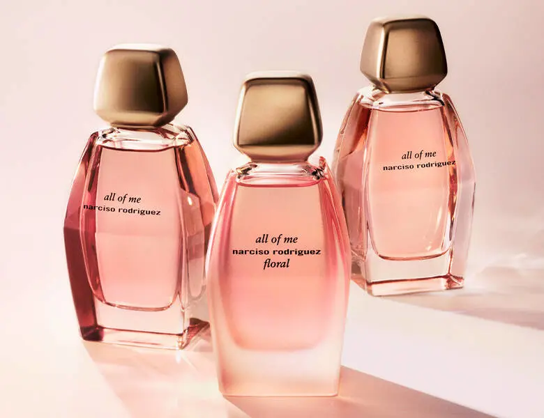 Her ses et billede med tre parfumeflakoner i rosa nuancer placeret side om side mod en lys baggrund. På flakonerne står teksten “all of me narciso rodriguez”, og på den midterste står også “floral”. Flakonerne har kantede former med gyldne låg.