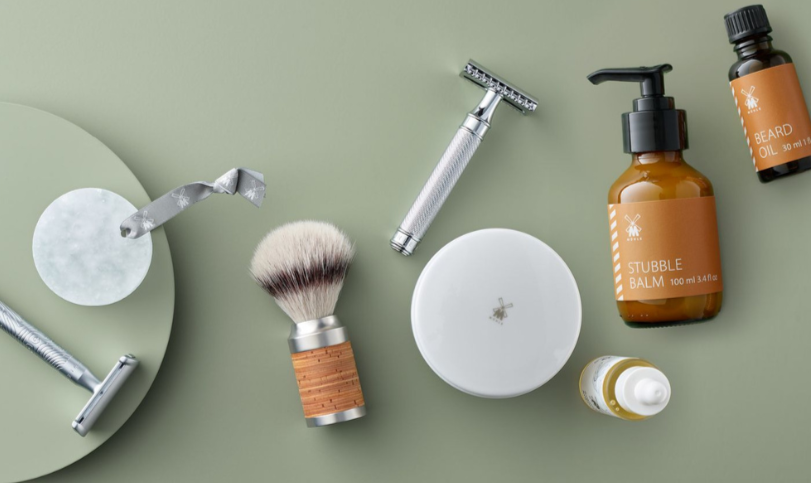 Mühle barberingsprodukter ligger arrangeret på lysegrøn baggrund, herunder skrabere, barberkost, skål, stubble balm og beard oil.