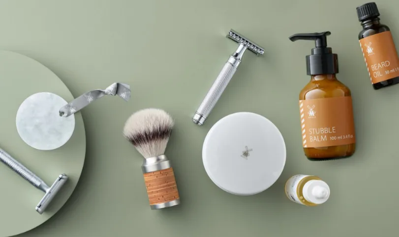 Mühle barberingsprodukter ligger arrangeret på lysegrøn baggrund, herunder skrabere, barberkost, skål, stubble balm og beard oil.