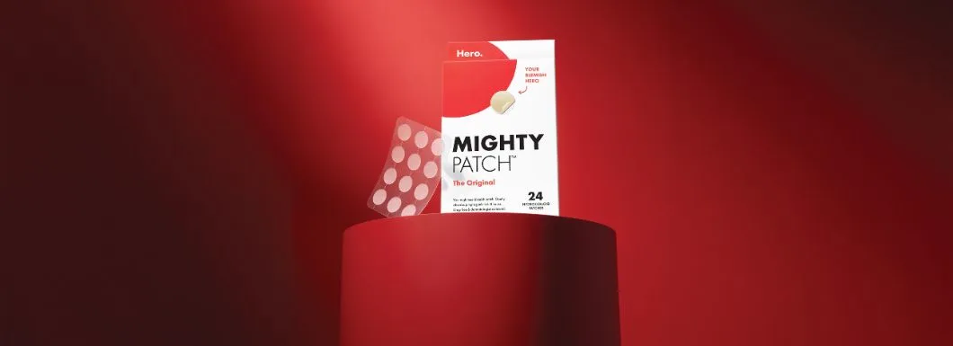Her ses en pakke Hero Mighty Patch The Original og et ark med plastre på en rød cylinder foran en mørkerød baggrund med et rødt lysfald ovenfra.