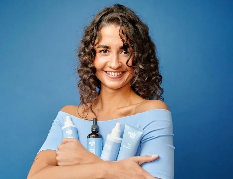 Her ses en kvinde med krøllet hår, der holder Yuaia Hair produkter i blå emballage på en blå baggrund.
