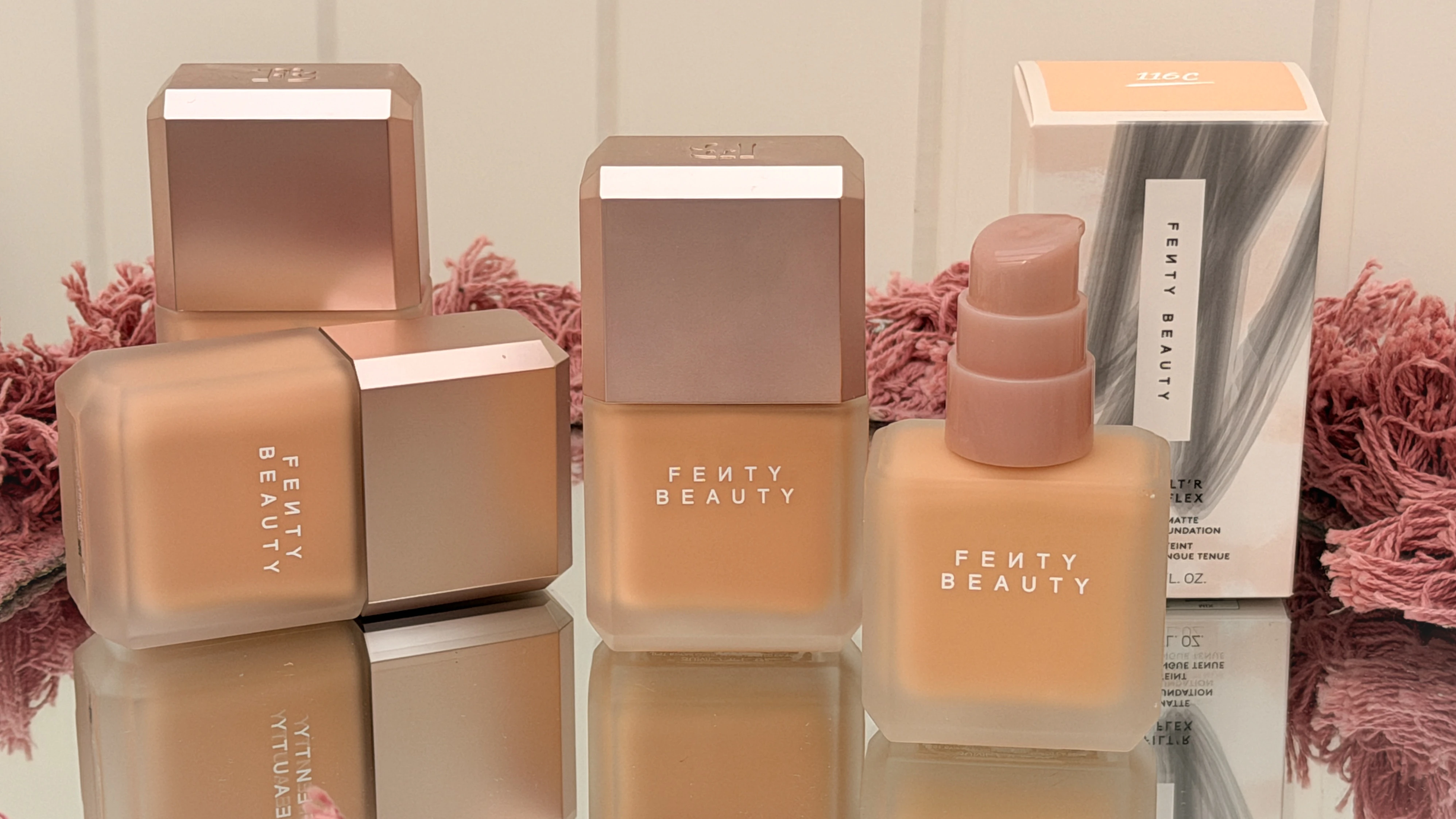 Tre Fenty Beauty foundationflasker og en æske med teksten 'Matte Foundation 116C'. De står på en reflekterende overflade.