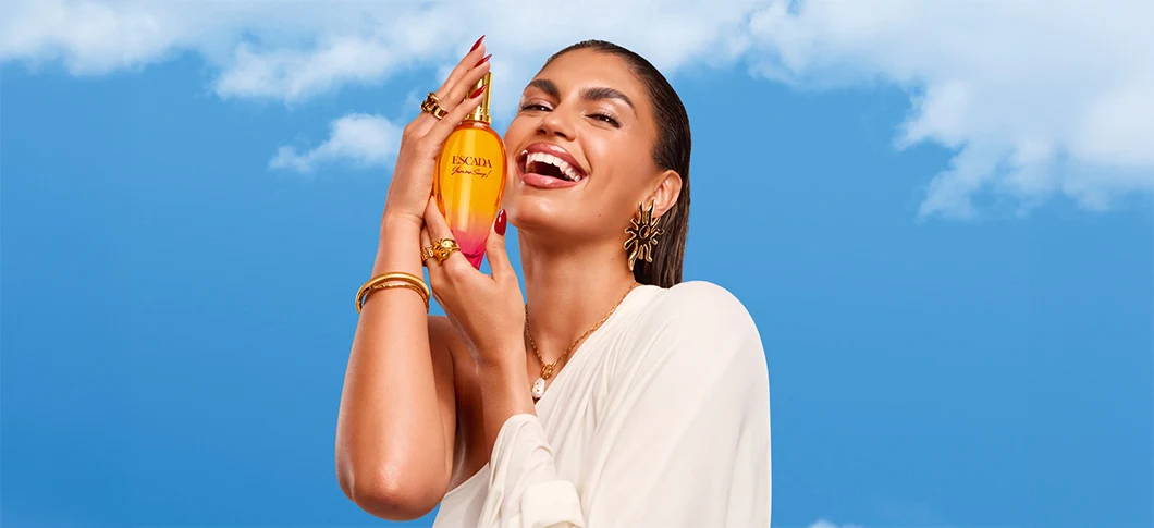 På billedet ses en smilende kvinde, der holder en orange Escada-parfume op mod en blå himmel.