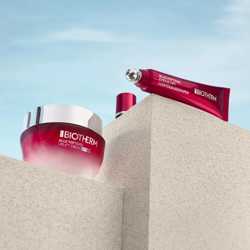 To Biotherm-produkter står placeret på lyse stenblokke mod en blå himmel. Til venstre ses en rød ansigtscreme med sølvfarvet låg og teksten “Blue Peptides Uplift Cream SPF 30”. Øverst til højre ligger en rød tube med metalapplikator og teksten “Blue Peptides Eyes & Lips Contour Reshaper”. Produkterne er fotograferet i et stilrent og eksklusivt miljø med fokus på hudpleje og anti-age pleje.