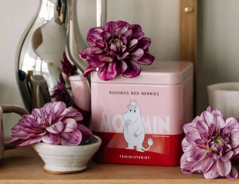 Her ses et rødt og lyserødt Moomin produkt blandt lilla blomster og kander.
