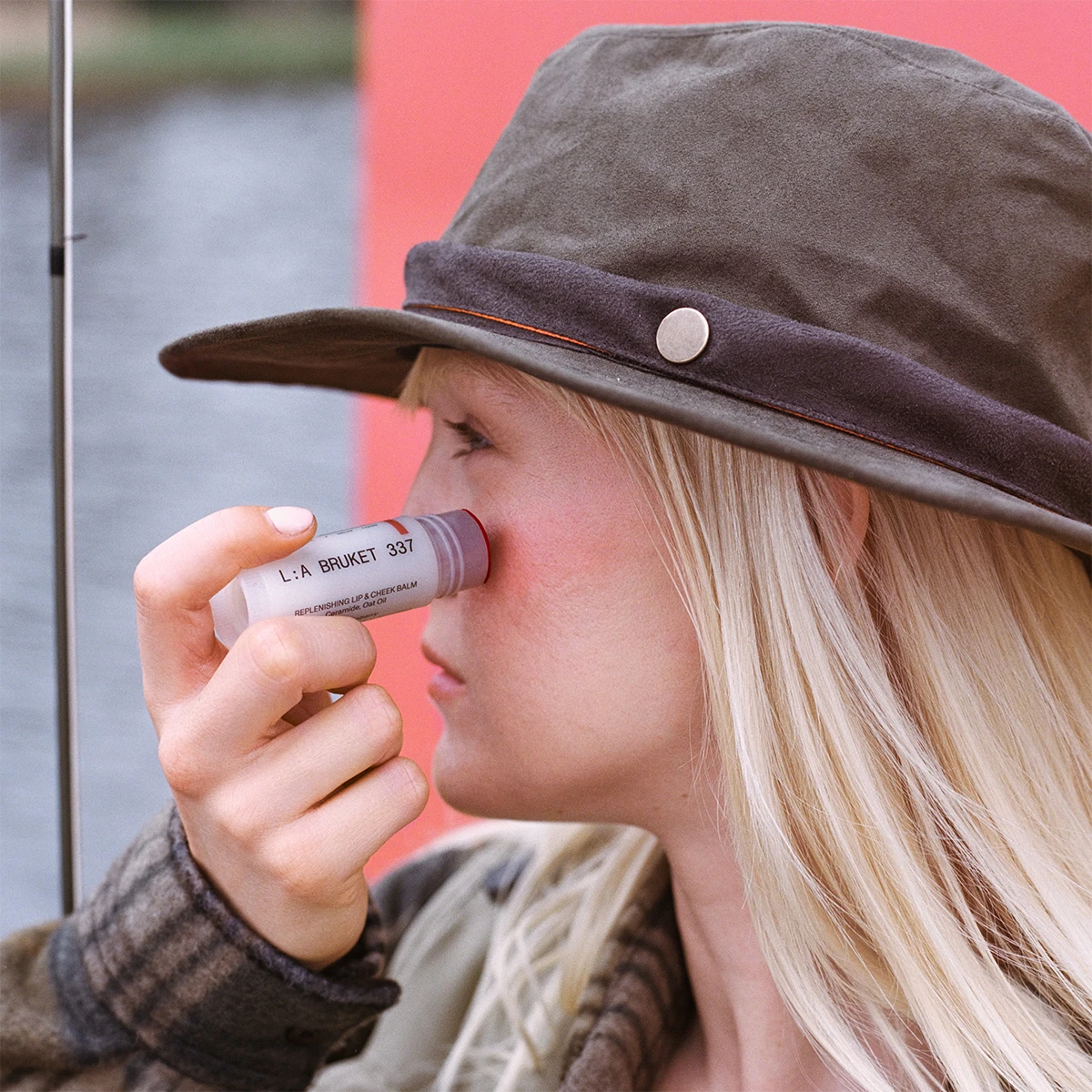 Person med blondt hår og hat påfører L:A BRUKET 337 Replenishing Lip & Cheek Balm på kinden.