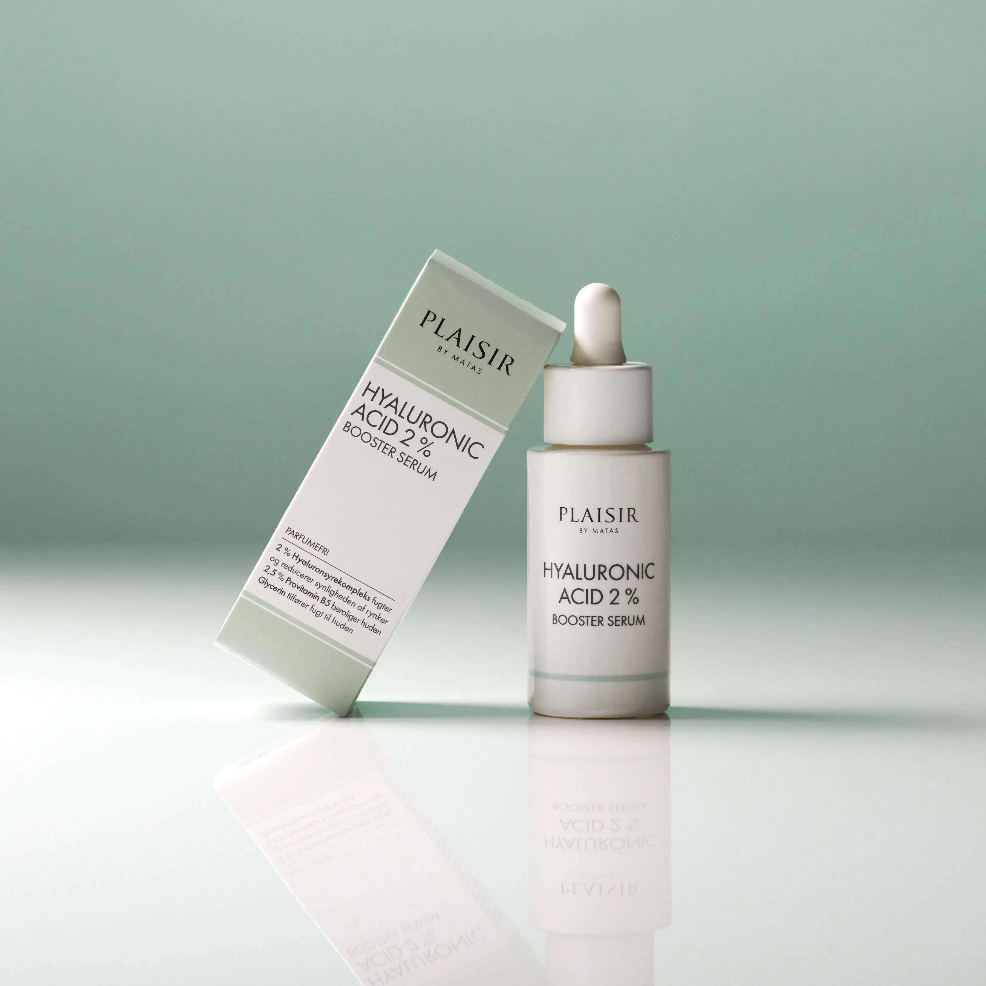 En flaske Plaisir Hyaluronic Acid 2% Booster Serum står på en blank overflade mod en grøn baggrund. Ved siden af står produktets æske let lænet. Flasken har pipette og spejler sig i underlaget.
