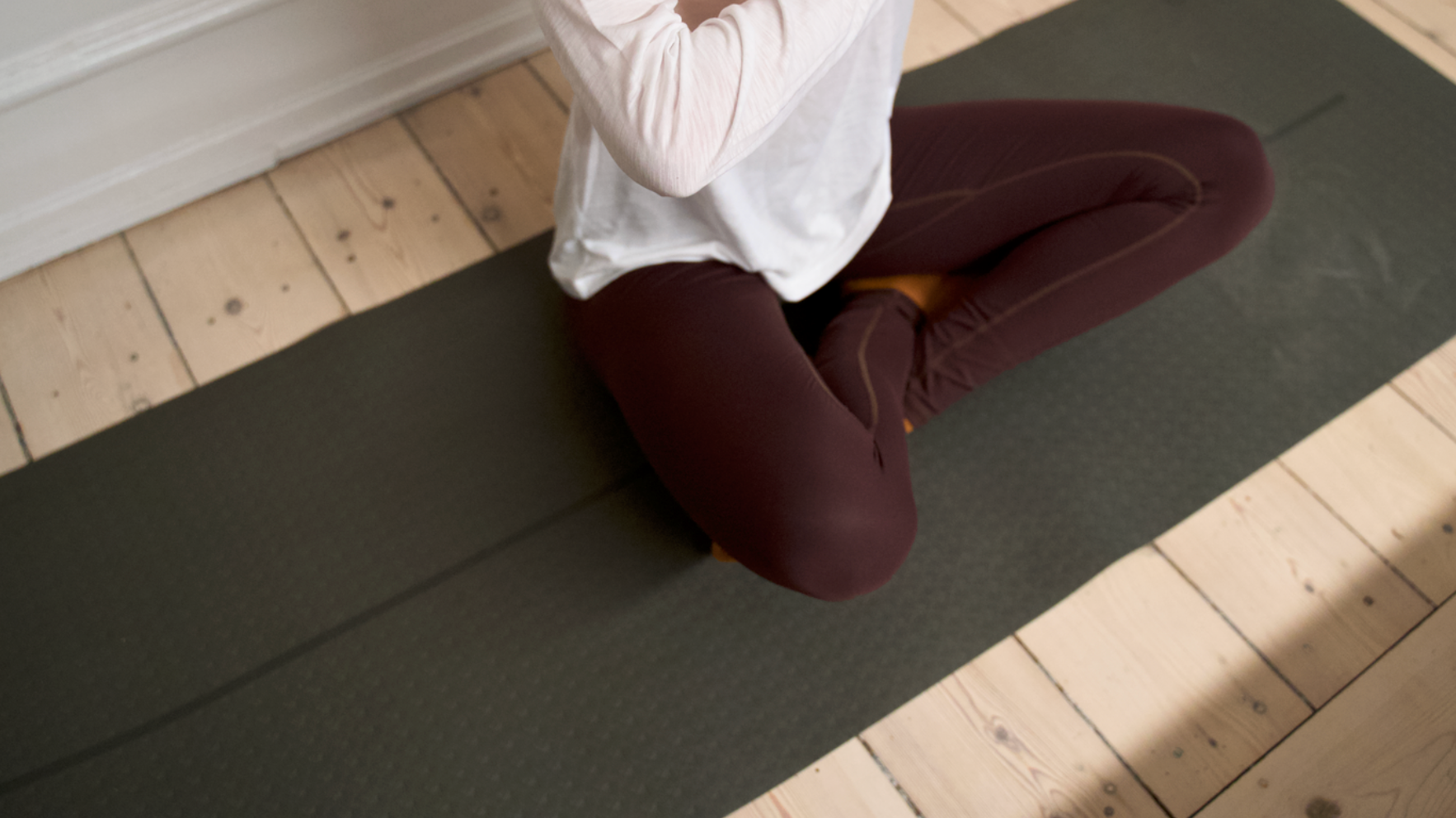 Person i meditationsstilling, siddende med krydsede ben på en mørk yogamåtte på et lyst trægulv.