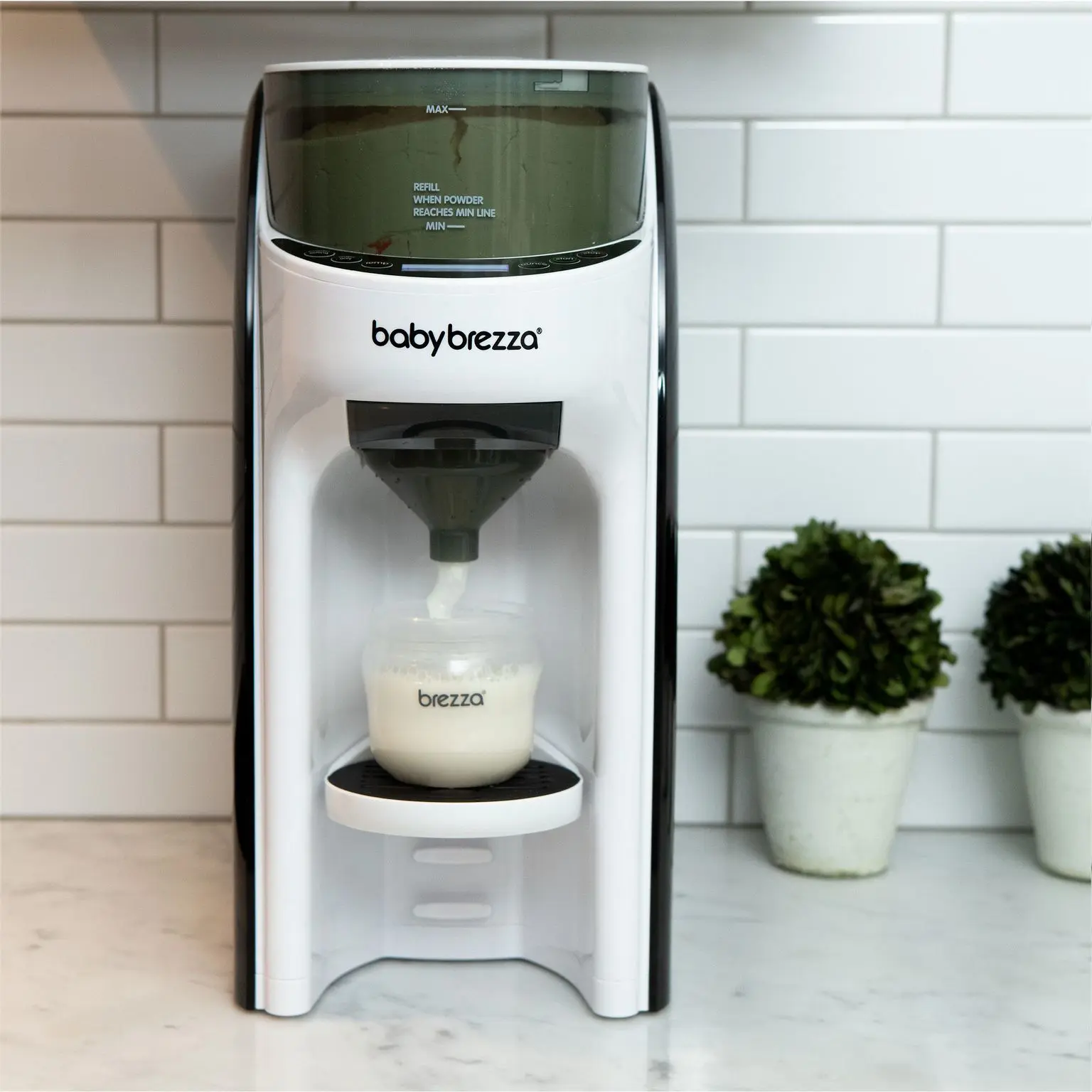 Babybrezza modermælkserstatningsmaskine fylder en flaske. Maskinen viser teksten 'babybrezza', 'MAX', 'MIN' og 'REFILL WHEN POWDER REACHES MIN LINE'. Flasken er mærket 'brezza'.