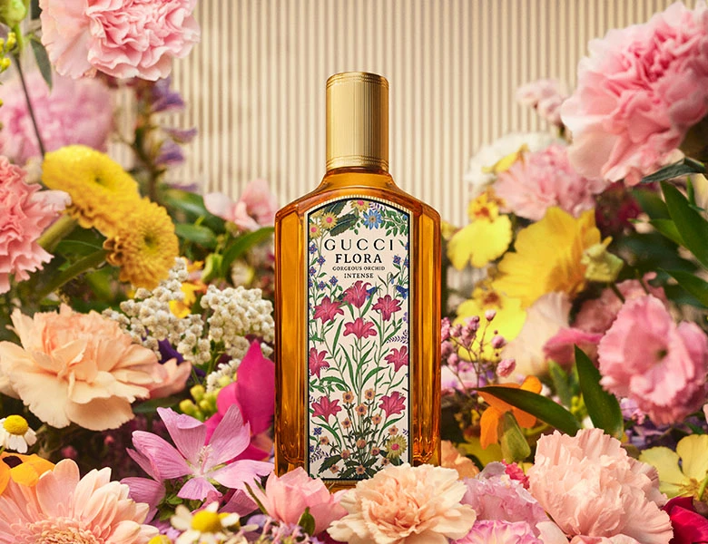 Her ses et billede med en parfumeflakon fra Gucci Flora placeret blandt farverige blomster mod en lys baggrund. Flakonen har en blomstret etiket og et gyldent låg og er omgivet af lyserøde, gule og hvide blomster.