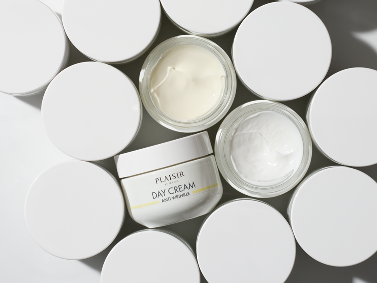 Flere hvide kosmetikbøtter, nogle med låg, og to åbne der viser creme. Én mærket 'Plaisir by Matas Day Cream Anti Wrinkle'.