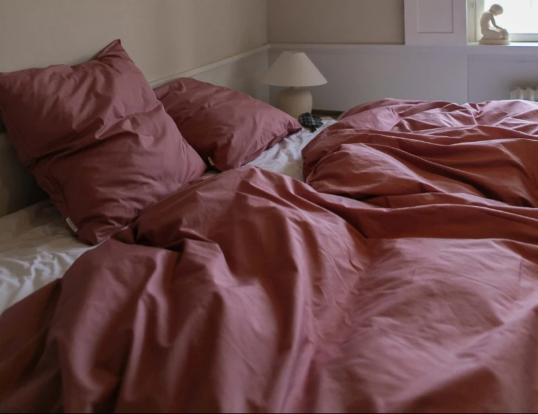 Her ses en seng redt med rosa sengetøj og puder i et lyst soveværelse.