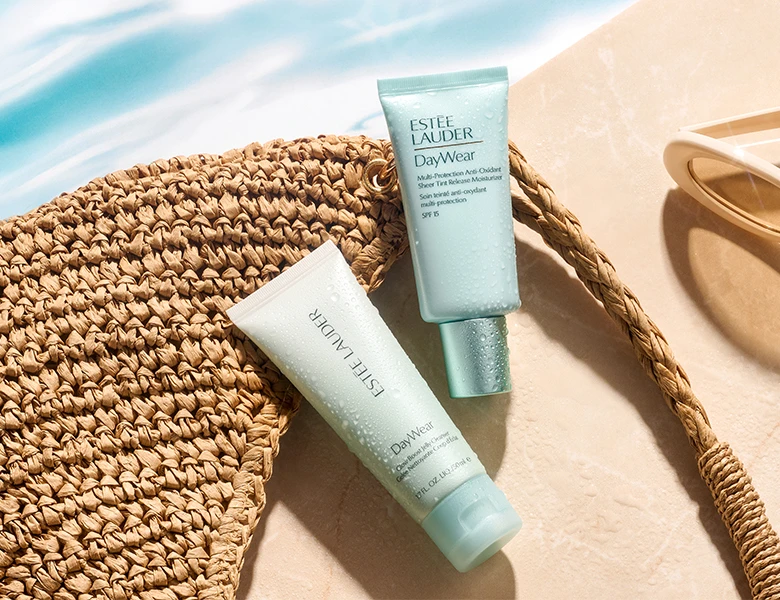 Her ses et billede med hudplejeprodukter fra Estée Lauder placeret ved en stråtaske og solbriller. På produkterne står teksten “DayWear”, og emballagen er i lyseblå nuancer.