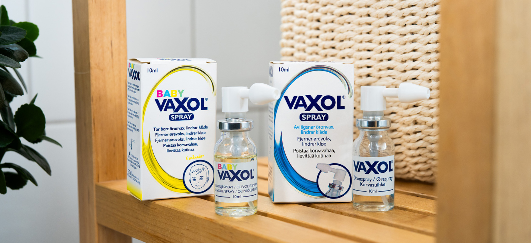 Baby Vaxol og Vaxol øresprays i æsker og små flasker står på en træhylde i et lyst badeværelsesmiljø med flettet kurv i baggrunden.