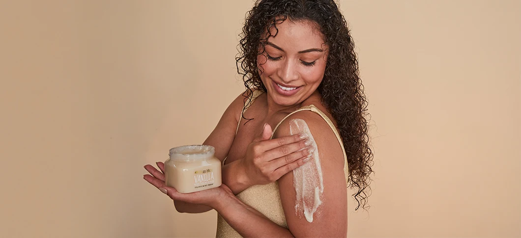 Her ses en kvinde, der smører body scrub på sin skulder og holder en krukke fra Tree Hut foran en beige baggrund.