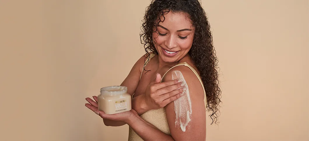 Her ses en kvinde, der smører body scrub på sin skulder og holder en krukke fra Tree Hut foran en beige baggrund.