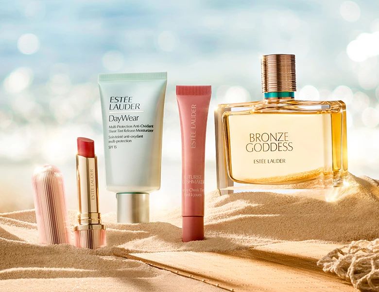 Her ses et billede med produkter fra Estée Lauder placeret i sand ved havet. Der ses blandt andet læbeprodukt, hudpleje og parfumen “Bronze Goddess”. Produkterne er placeret i et sommerinspireret miljø med strand og vand i baggrunden.