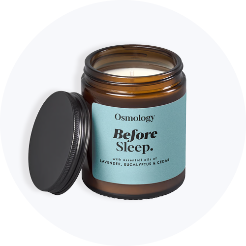Brun glas krukke med lys og åbent sort låg. Etiketten siger 'Osmology Before Sleep. with essential oils of LAVENDER, EUCALYPTUS & CEDAR'.