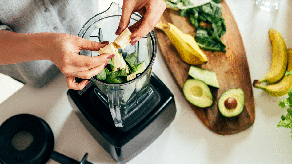 Her ses hænder, der lægger banan og grøntsager i en blender ved siden af avocado og frugt på et køkkenbord.