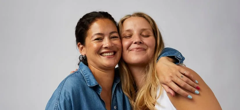 To kvinder krammer tæt og smiler foran en lys grå baggrund. Kvinden til venstre har mørkt hår sat op og en blå denimskjorte på. Hun smiler direkte mod kameraet. Kvinden til højre har langt blondt hår og en hvid top på. Hun har lukkede øjne og læner sig ind mod den anden kvinde med armen omkring hendes skuldre. Begge ser glade og afslappede ud.