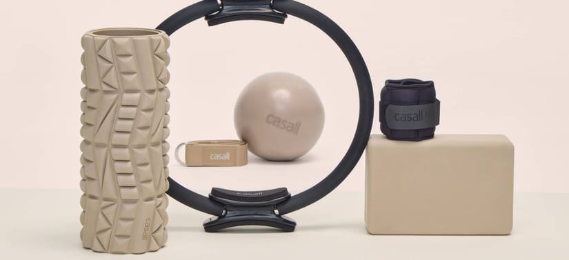 Her ses en beige træningsmåtte, en beige foam roller på højkant, en lyserød hvid bold og en beige yoga band i baggrunden igennem en yoga ring i midten, og til højre en sort håndledsvægt ovenpå en beige yoga brick. Alt foran en lyserøde væg