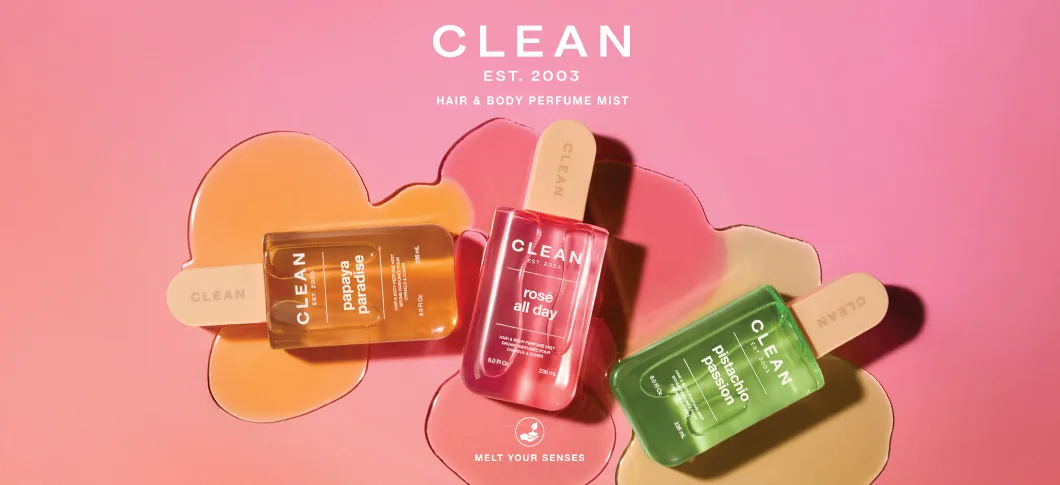 Her ses et billede med tre produkter fra CLEAN placeret mod en lyserød baggrund med farvede væskeformer. Øverst står teksten “CLEAN EST. 2003 HAIR & BODY PERFUME MIST”, og nederst står “MELT YOUR SENSES”. På produkterne står blandt andet “papaya paradise”, “rosé all day” og “pistachio passion”.