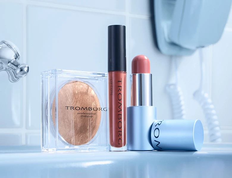 Tre Tromborg makeup-produkter: en gylden-bronze pudder, en Lip Cute lipgloss og en læbestift i en lys koralfarve.