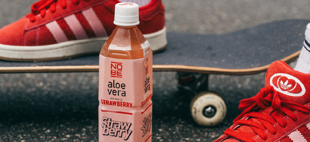 På billedet ses NOBE Strawberry på astfalt foran fødder med røde sneakers hvor den højre fod står på skateboard 