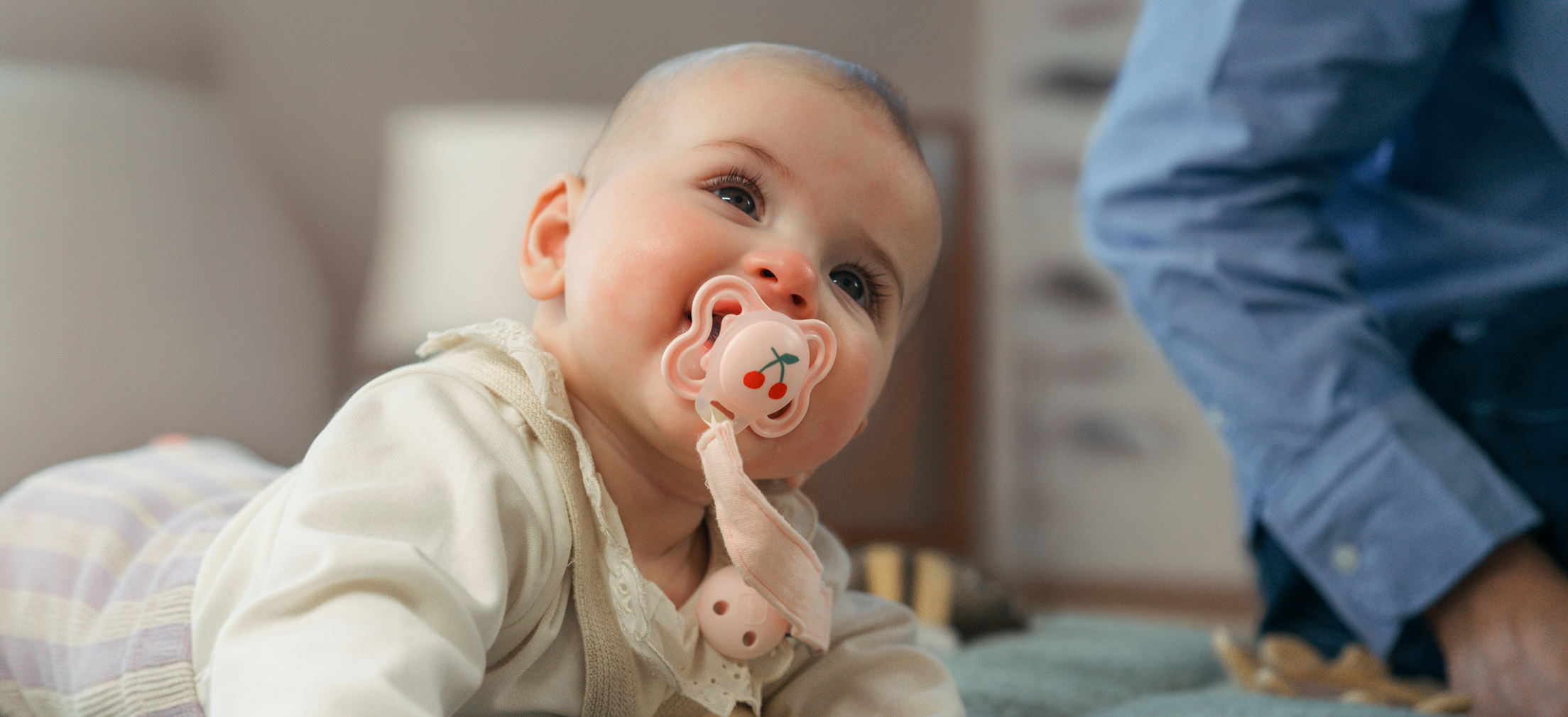 Baby ligger på maven og kigger op med lyserød sut i munden, mens en voksen sidder ved siden af i blødt, hjemligt miljø.