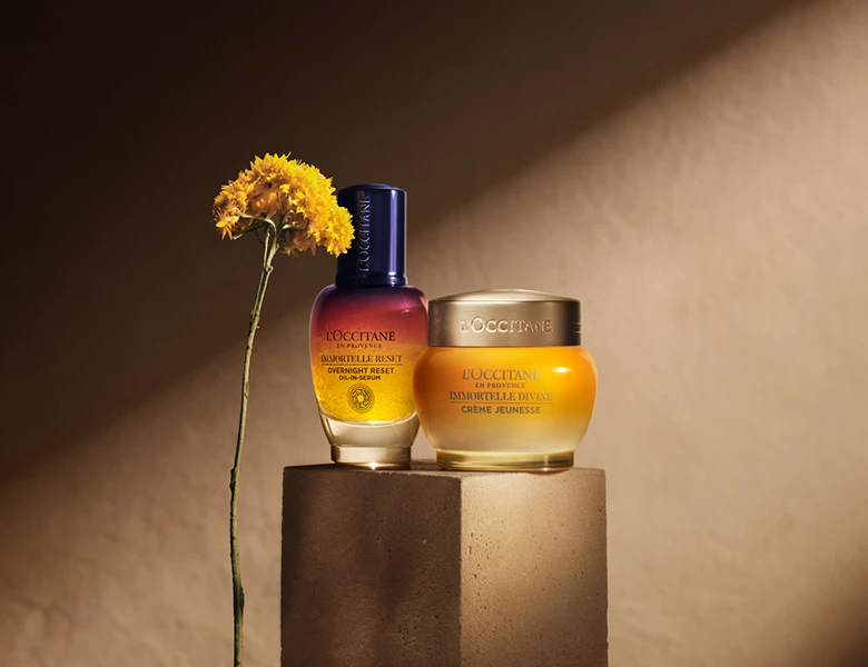 Her ses L’Occitane hudplejeprodukter – serum og ansigtscreme ved siden af en gul blomst på en varm beige baggrund.
