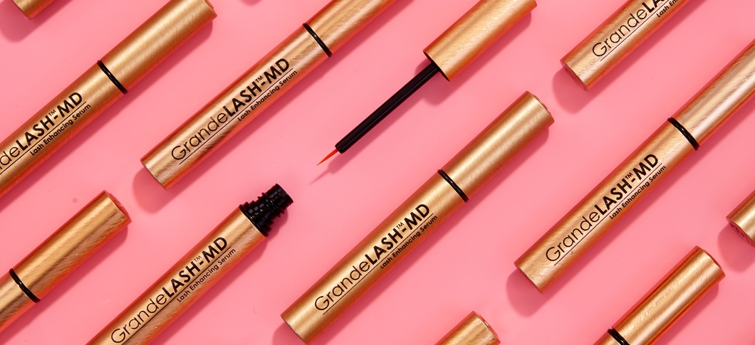 Her ses et billede med flere produkter fra GrandeLASH-MD placeret diagonalt mod en lyserød baggrund. På produkterne står teksten “GrandeLASH-MD Lash Enhancing Serum”, og nogle af dem er åbne med synlig applikator.