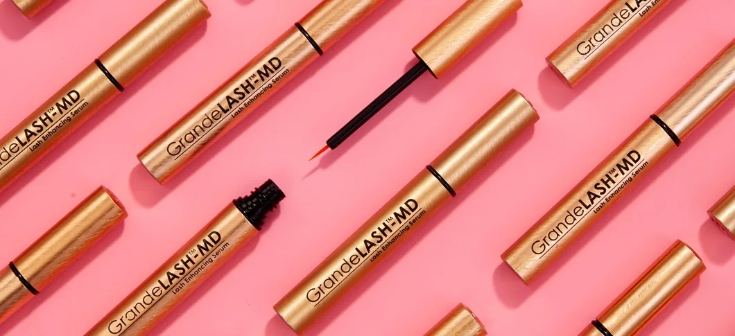 Her ses et billede med flere produkter fra GrandeLASH-MD placeret diagonalt mod en lyserød baggrund. På produkterne står teksten “GrandeLASH-MD Lash Enhancing Serum”, og nogle af dem er åbne med synlig applikator.