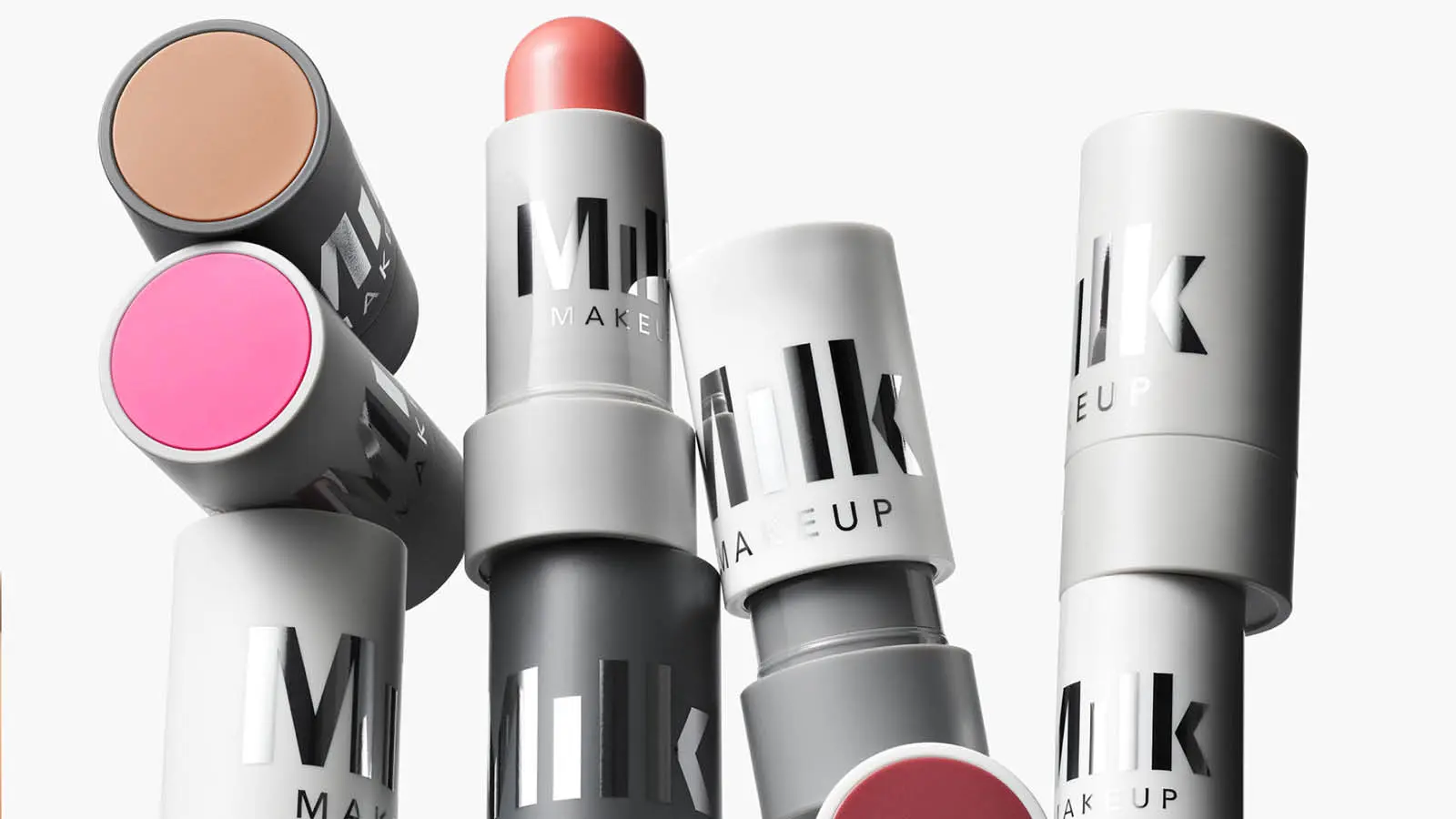 Her ses et udvalg af Milk Makeup-produkter i stickformat med grå emballage. Flere produkter er åbne, så nuancer i beige, pink, koral og bordeaux er synlige. Produkterne er arrangeret tæt sammen mod en lys baggrund.