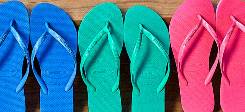 Her ses tre par Havaianas klipklapper - et par i blå, et par i tyrkisgrønne og et par i pink. De står på et mørkt trægulv.