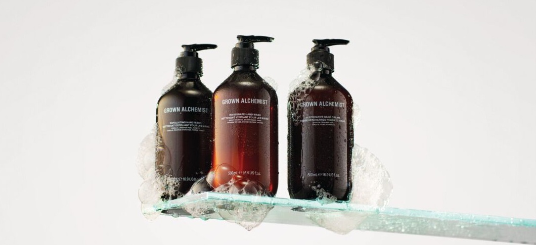 Her ses et billede med tre produkter fra Grown Alchemist placeret på en glashylde mod en lys baggrund. På produkterne står teksten “GROWN ALCHEMIST”. Produkterne er i mørke flasker med pumpe og er omgivet af skum.