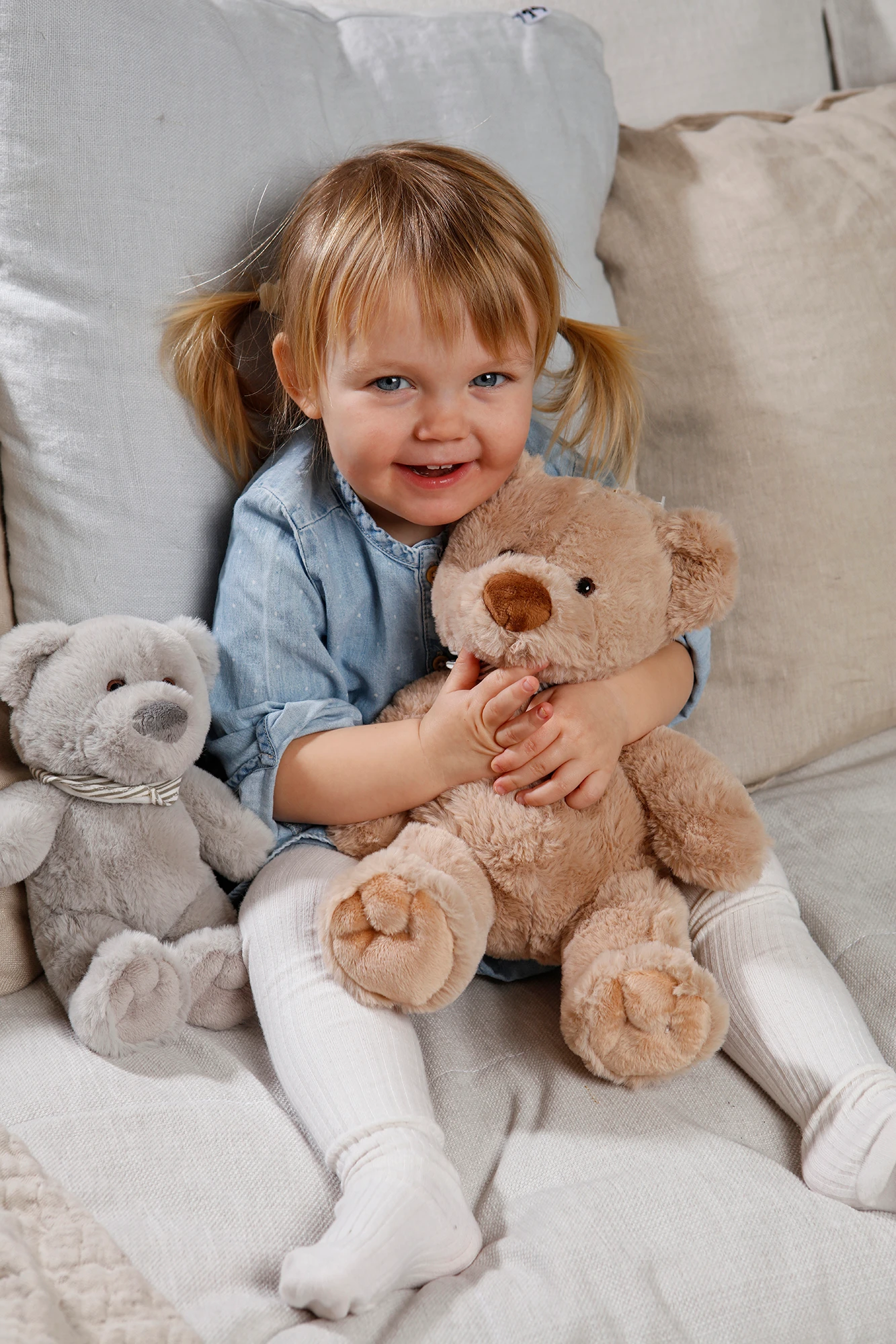 Smilende barn med blonde hestehaler holder en brun bamse, en grå bamse sidder ved siden af.