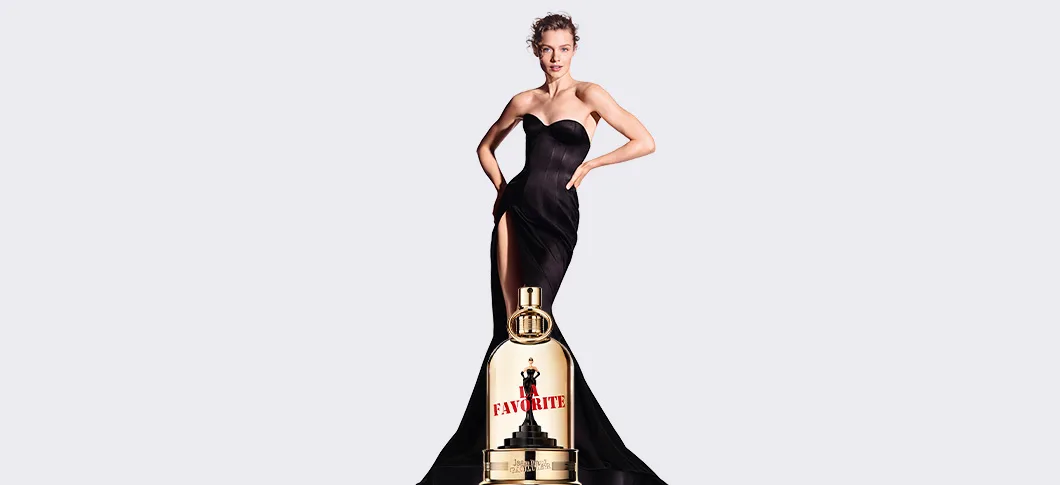 Her ses et billede af en person iført en lang, sort kjole mod en lys baggrund. Foran personen ses en parfumeflakon med teksten “LA FAVORITE” fra Jean Paul Gaultier, som er placeret centralt i billedet.