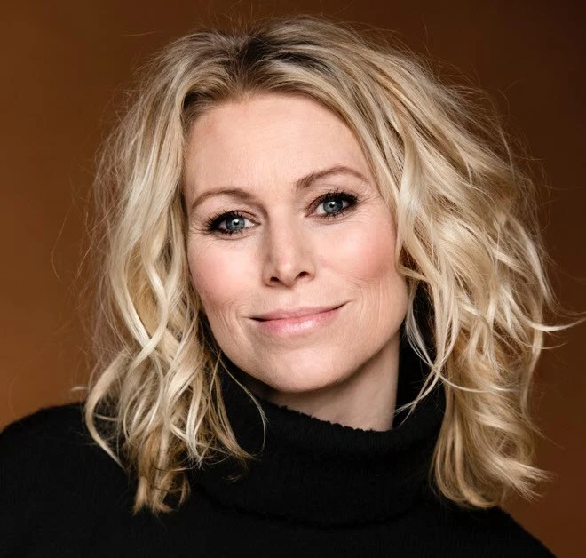 Portræt af Anna Bjerre: blond kvinde med bølget, skulderlangt hår, blå øjne og sort rullekrave mod en varm brun baggrund.